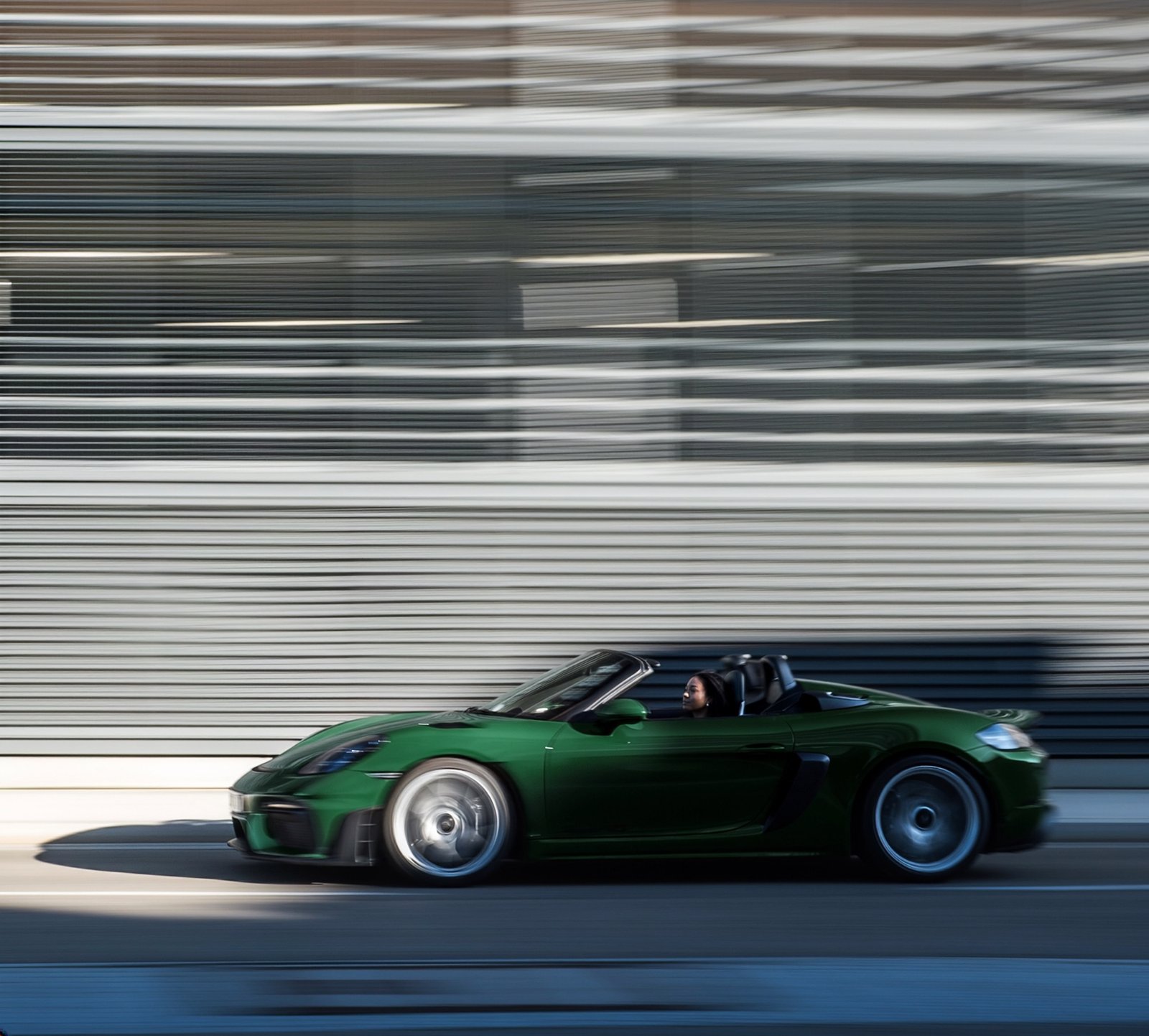 A green Porsche roadster driving past a striped building, shot with motion blur